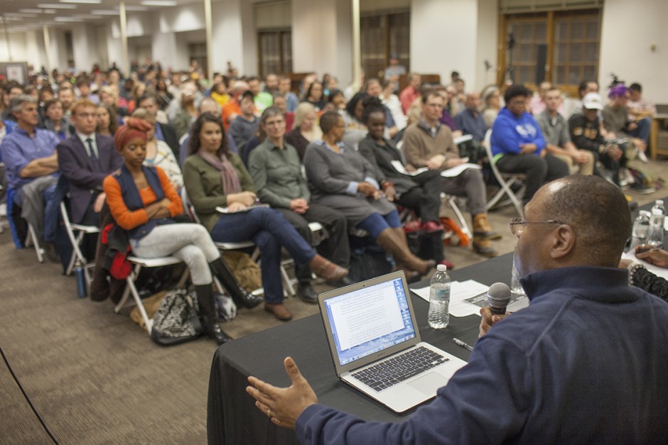 Crowd of people in chairs, facing a person speaking.