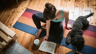 Young woman sitting on the carpet with her pet reading book and practicing yoga at the same time