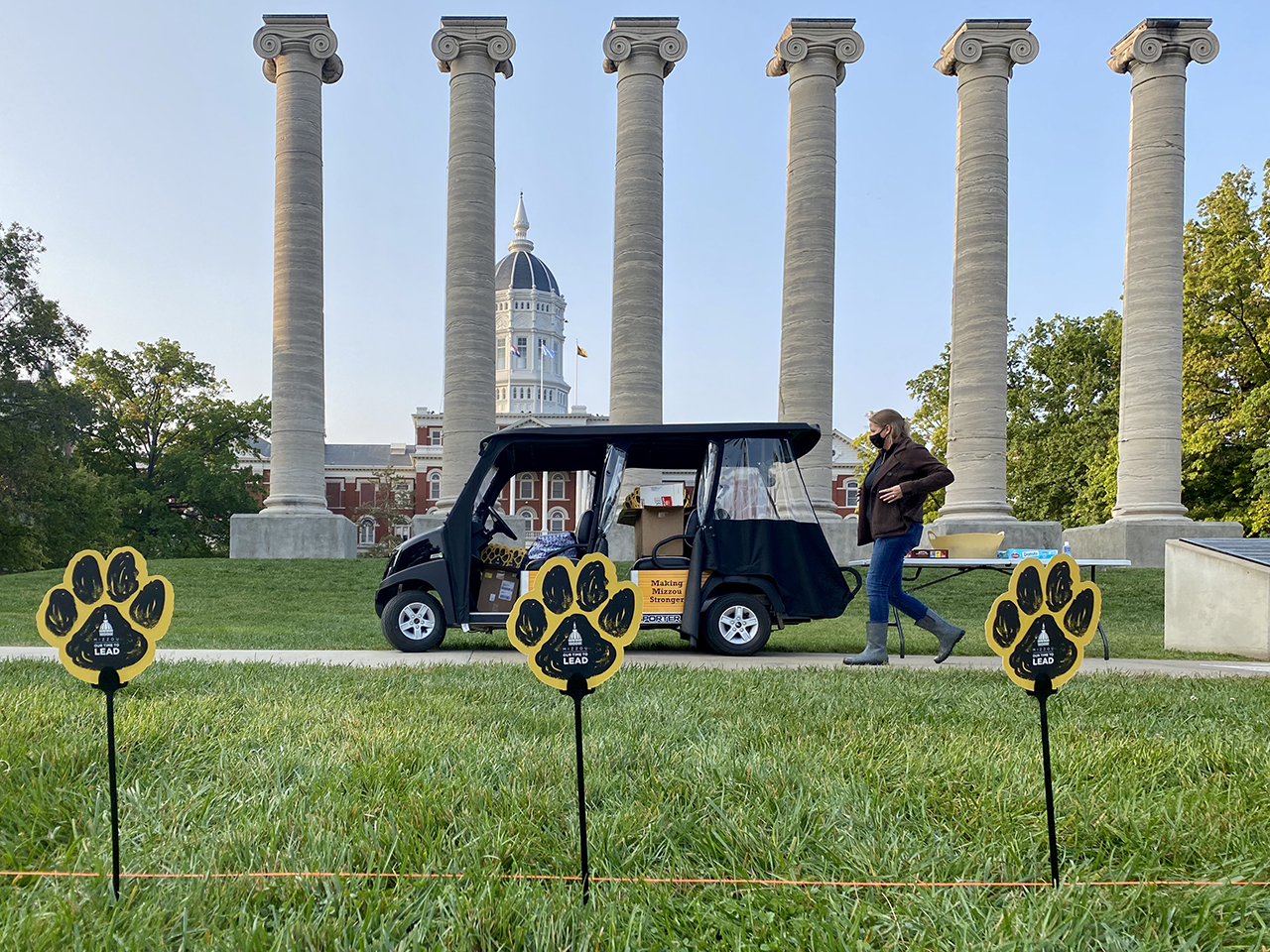 a woman walks in front of a golf cart with the columns in the background and paws on sticks in the foreground