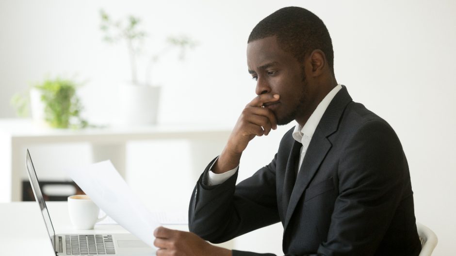 Black man sitting at a desk with a laptop, looking at a piece of paper