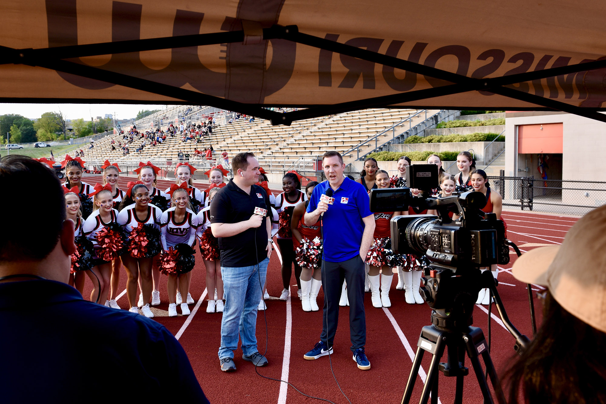two people talk on camera in front of a bunch of cheerleaders
