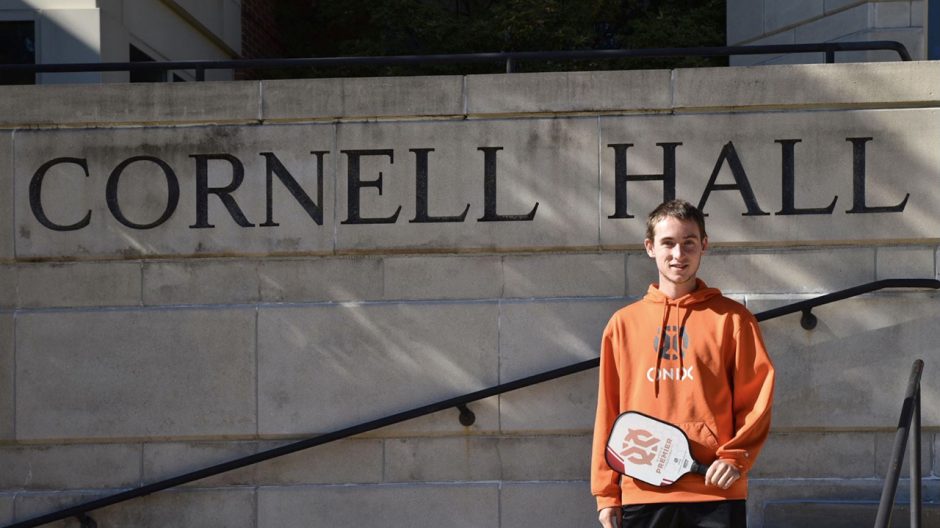 Dylan Frazier stands in front of Cornell Hall holding his pickleball paddle