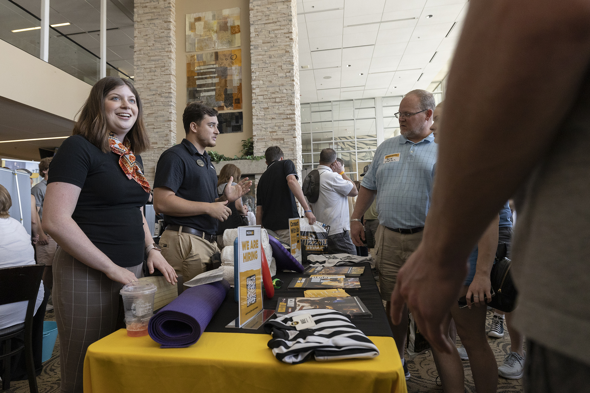 a woman smiles as a man interacts with a student and parent at an indoor fair