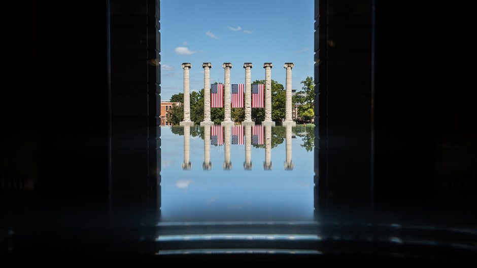 a reflection of the columns through the window of jesse hall. the columns have the american flag hanging between them