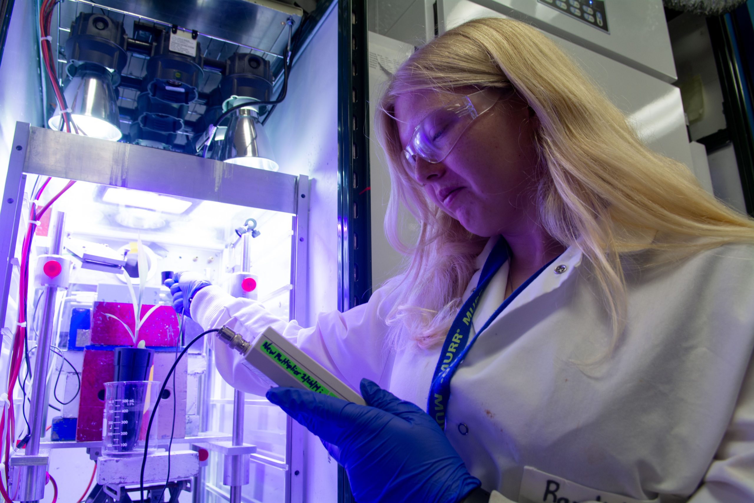 a woman checks a scientific instrument in front of a plant growing in a box in a scientific lab