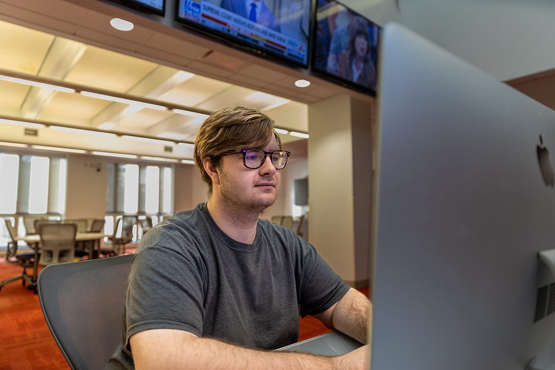 Brandon Ford types on a computer in front of bank of TVs in the Reynolds Journalism Institute