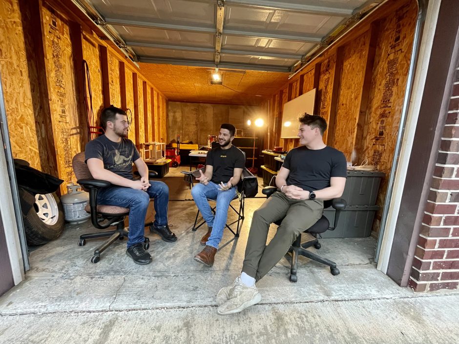 three men sit in a garage and talk and smile