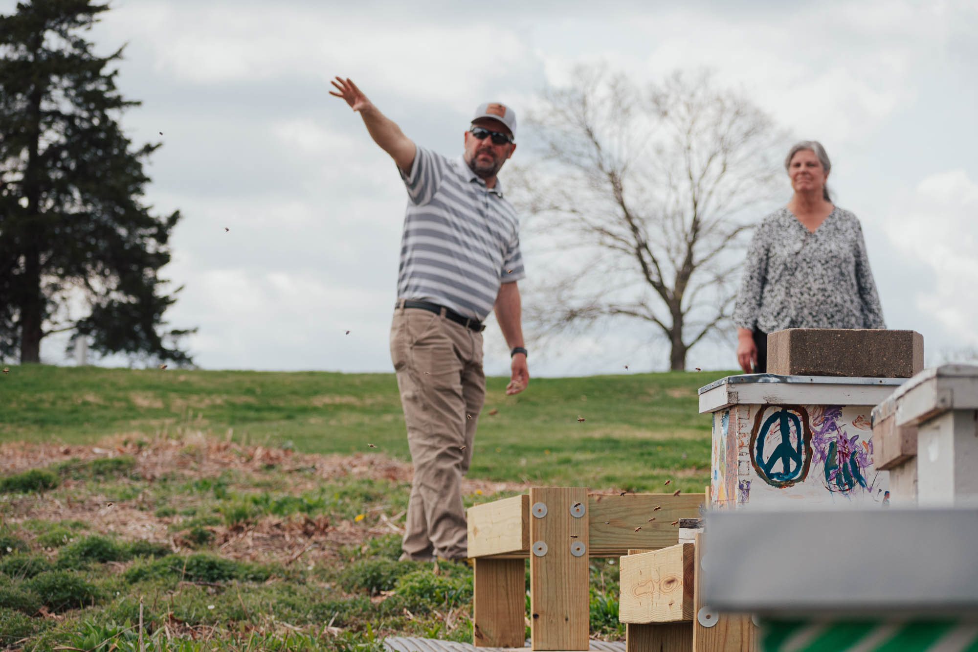 a man gestures to the left. there are beehives in front of him. a woman looks on.