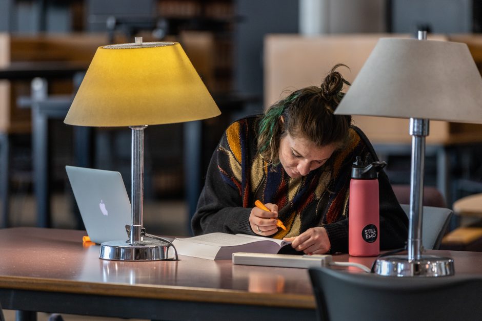 Female student at table in a library with laptop open who is actively highlighting text in a book next to a pink water bottle 