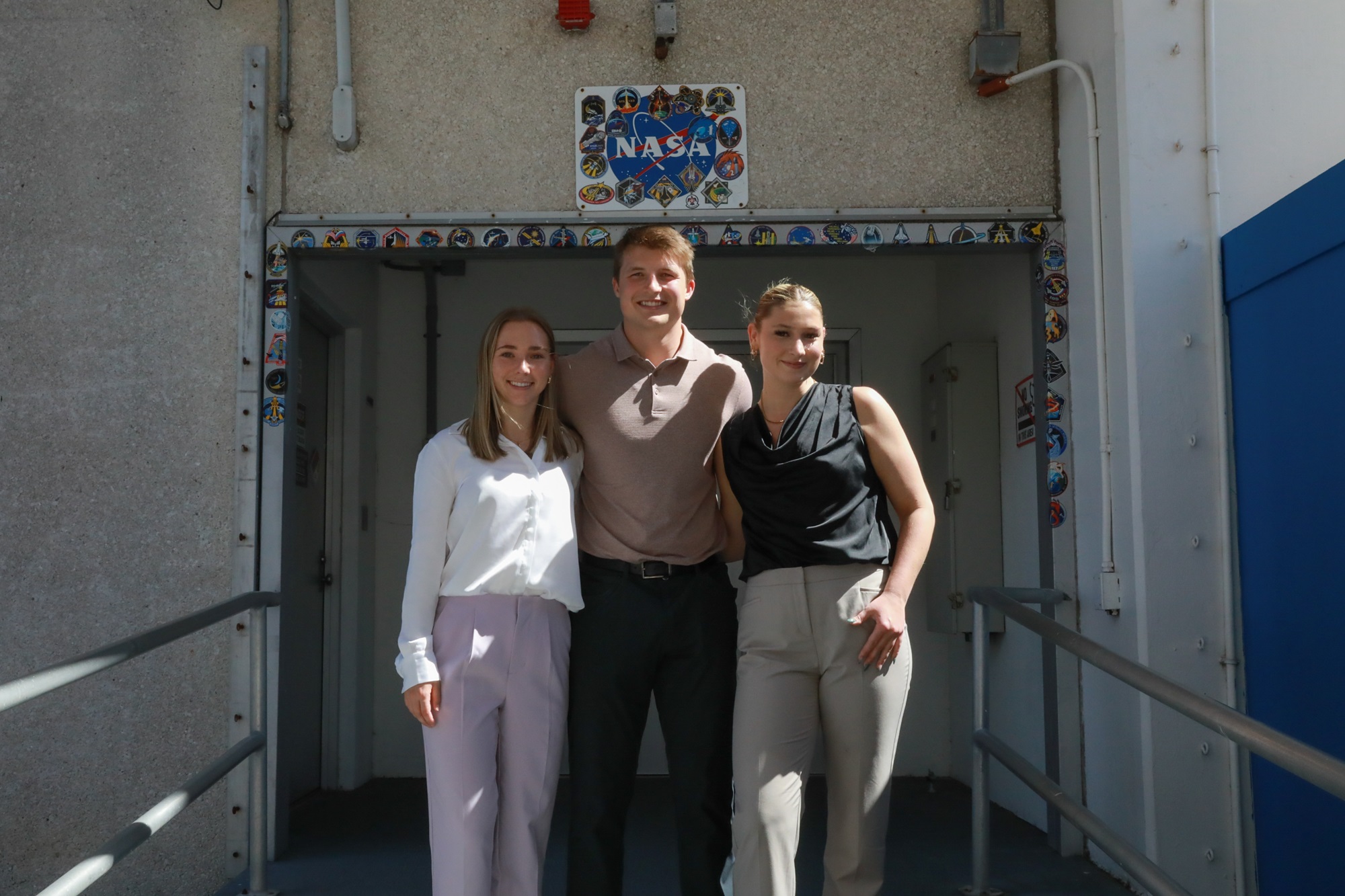 three people standing in front of the Kennedy Space Center’s astronaut walkout doors.