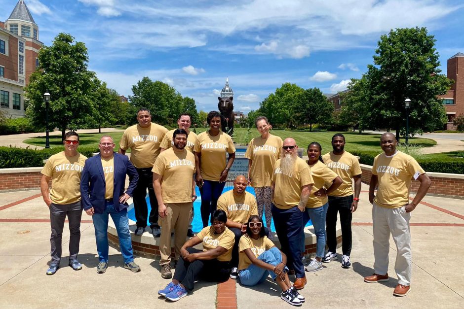 A group of veterans in gold Mizzou shirts stand in front of a fountain.