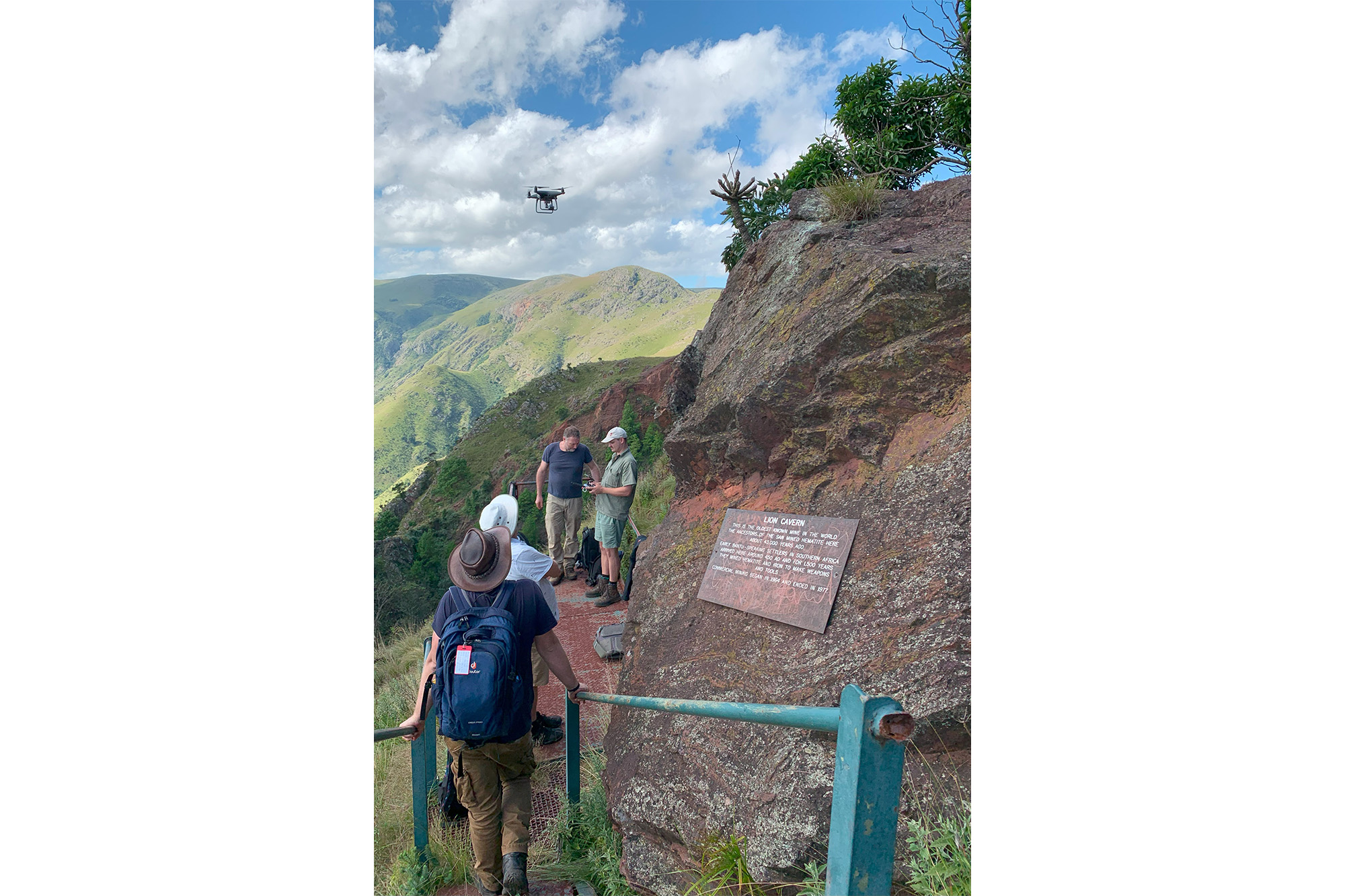 Several people walking along a mountainside trail with a drone overhead