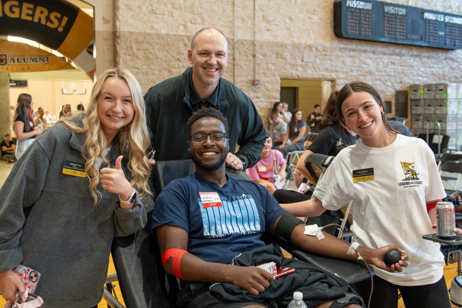 Four people smile at camera as one gives blood