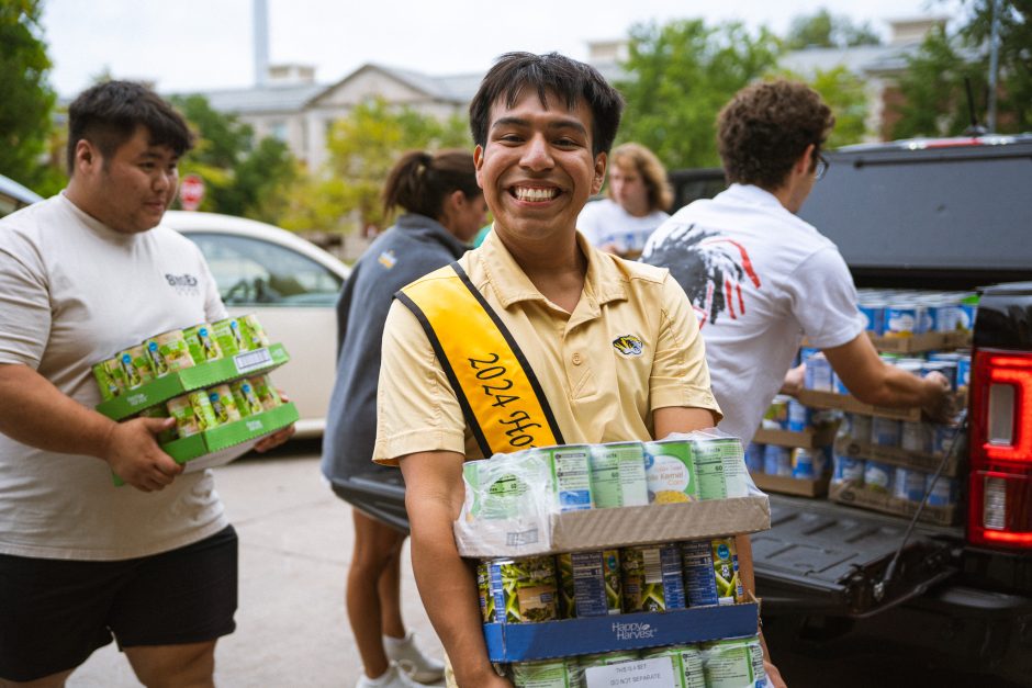 Student smiles at camera holding box of canned goods