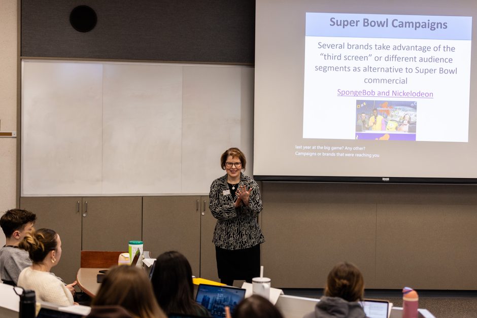 Professor in front of screen speaks to students.