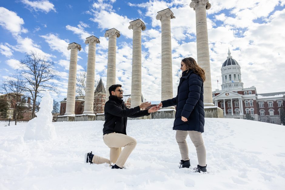 A man (left) kneels in the snow proposing to a woman (right) while holding her hand. They're in front of the columns and Jesse Hall. The day is bright with clouds rolling in.