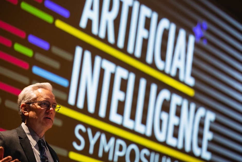 A man is seen in the lower left hand corner of the frame, speaking to an audience (unseen).  The rest of the frame is a black slide deck presentation with the words "Artificial Intelligence Symposium" and some colored lines for decoration.