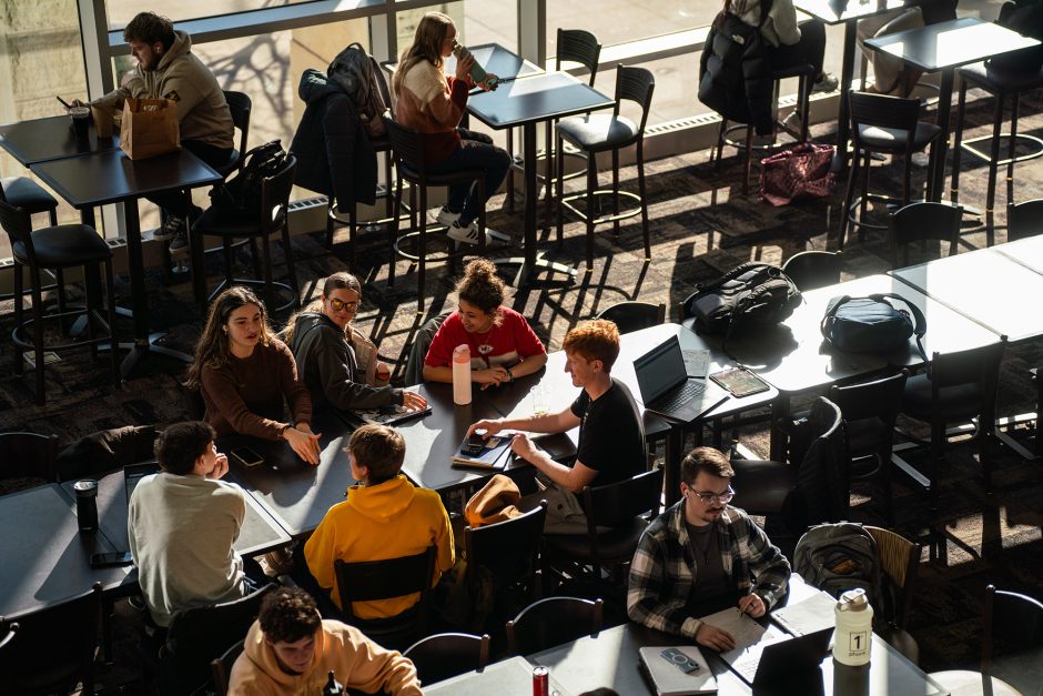 A bird's eye view of several long and short tables with students sitting at them. The students on the edges of the frame are on computers, notebooks or eating meals. The students in the center are in a lively discussion. Afternoon light fills in from the windows and casts long shadows. 