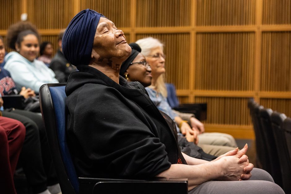 An older woman with vitiligo sits in an auditorium seat and has leaned back her head and closed her eyes. She's wearing a black zip up, hooded jacket and a navy head covering.