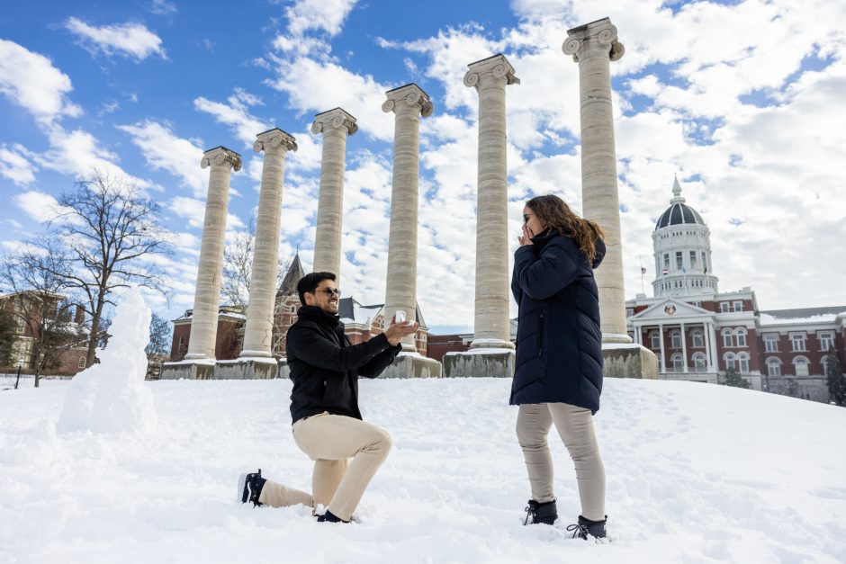 Person on one knee proposing in the snow in front of the Columns