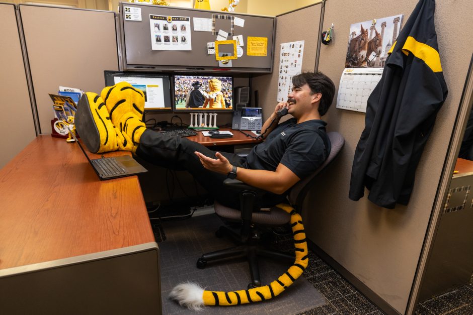 Person at a desk with Truman the Tiger feet propped up and wearing a tiger tail