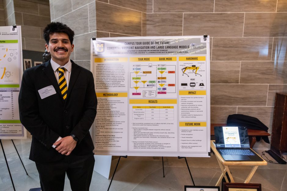 Student wearing black suit standing next to research poster