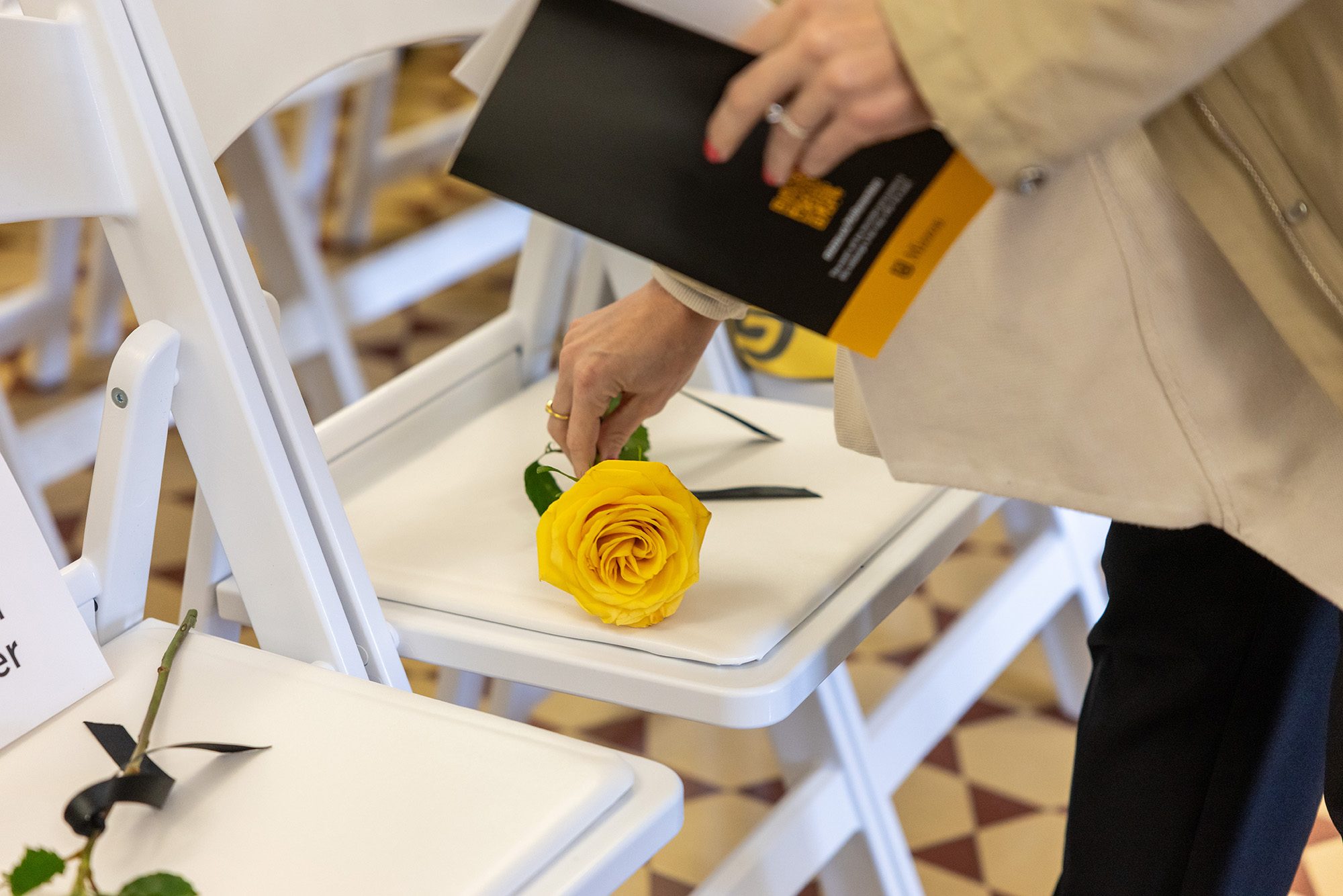 20260410_OP_MU-Remembers-025 A person's hand placing a yellow rose on a white chair