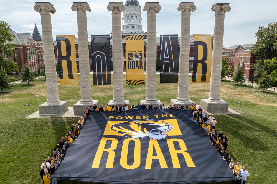 People holding a "Power the Roar" banner in front of the Columns