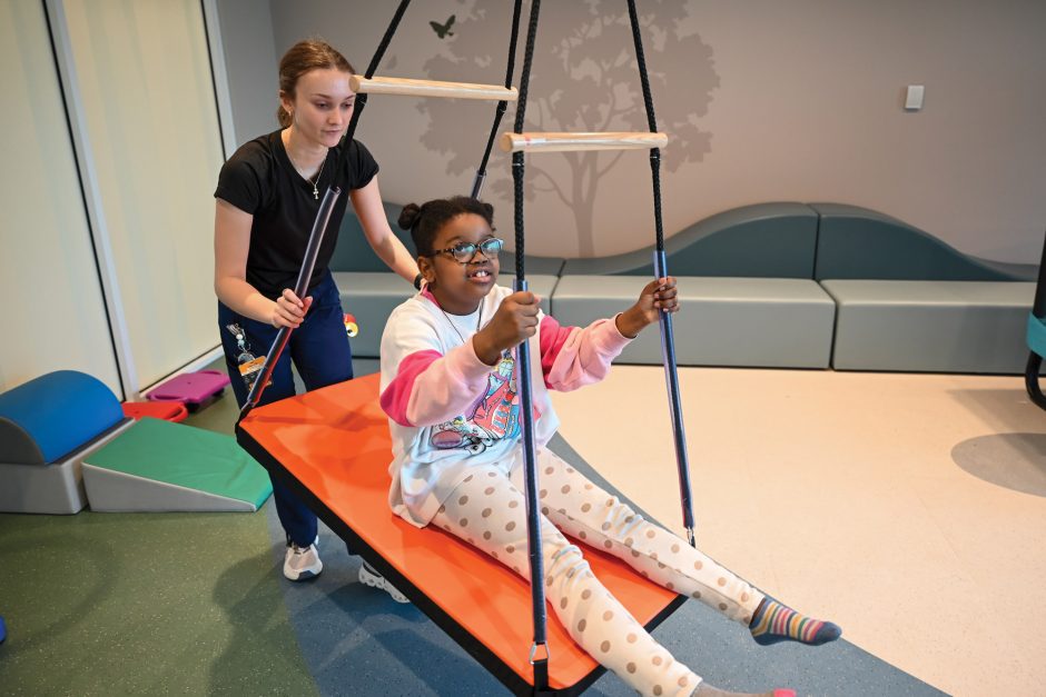 a young girl on an indoor swing set is pushed by a Thompson Center staffer
