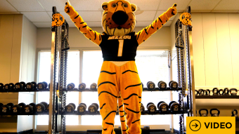 Truman the Tiger with his arms raised in a gym with the word video in the bottom right