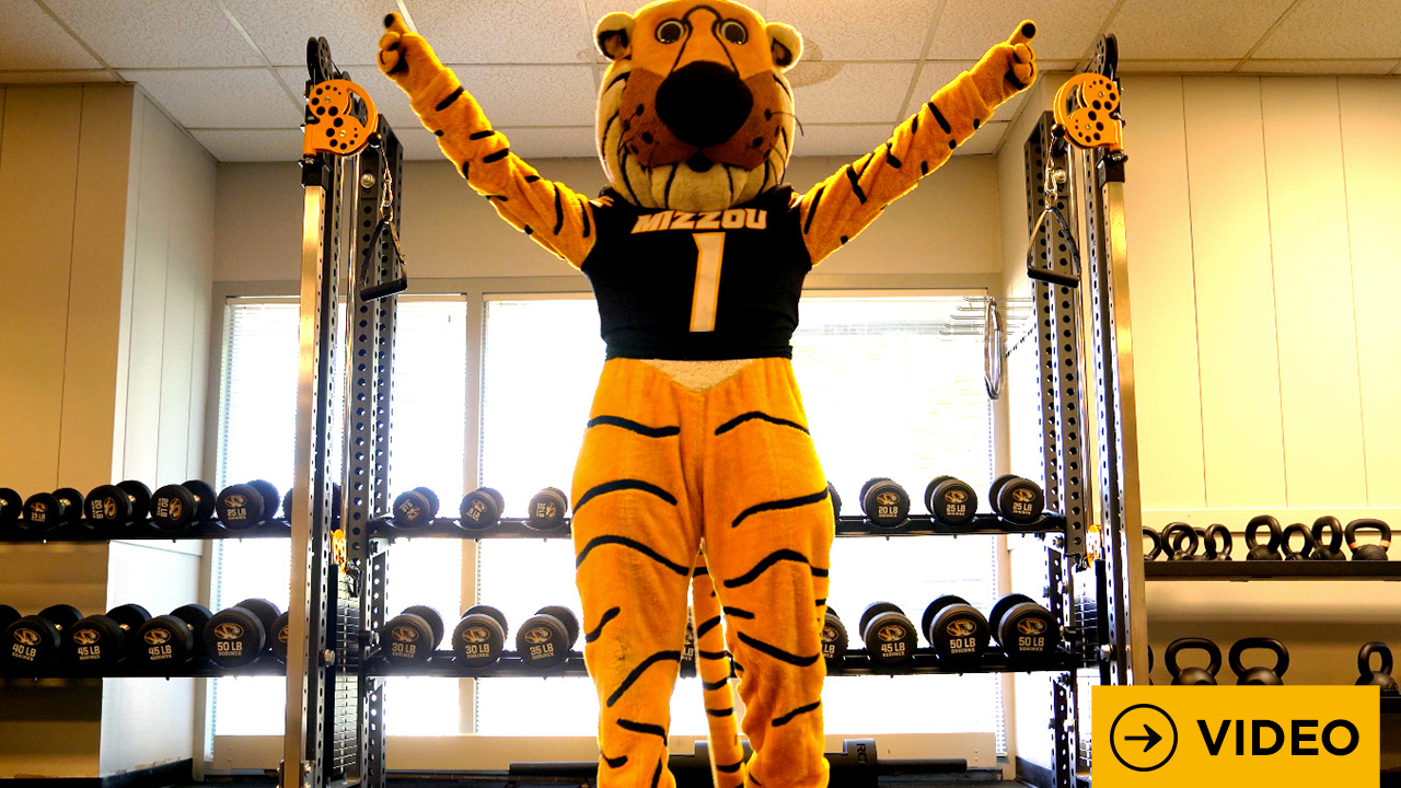 Truman the Tiger with his arms raised in a gym with the word video in the bottom right