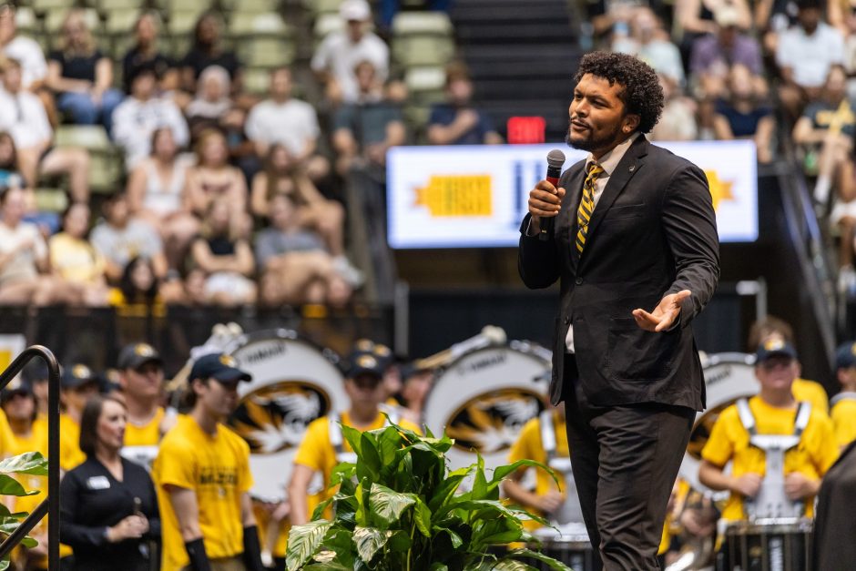 Tyson Ellison speaks on a stage on the floor of Mizzou Arena with Marching Mizzou and a crowd of students behind him