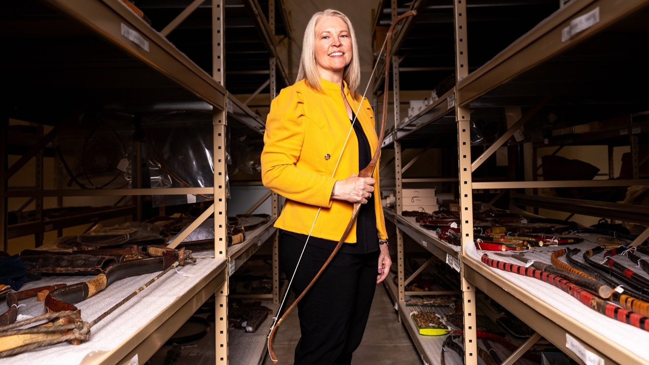 Candace Sall stands with her competition bow among shelves of archery artifacts that make up Mizzou's Grayson Archery Collection