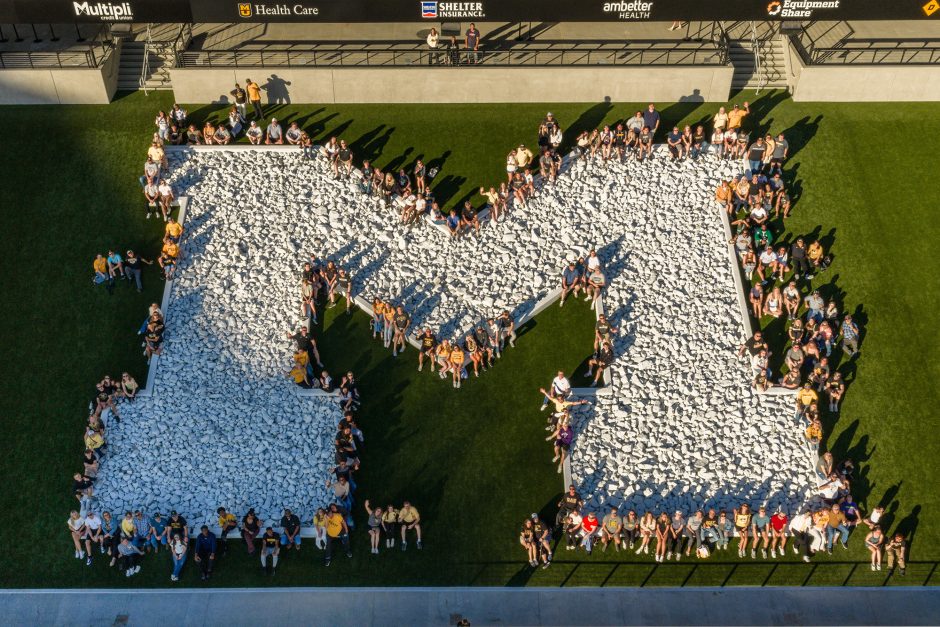 Aerial view of people surrounding a large white letter M on the ground